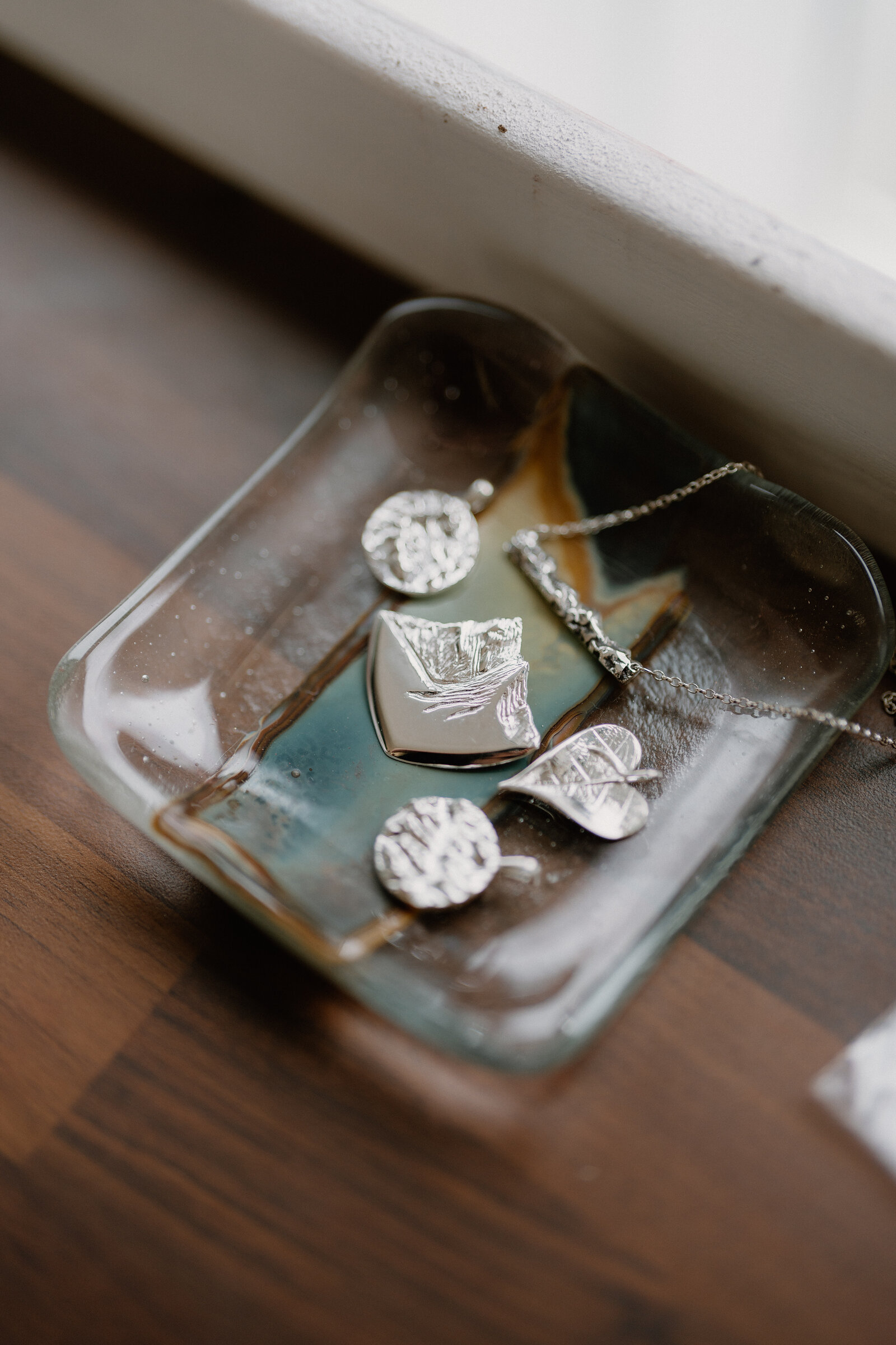 Silver pendant, disc earrings and a leaf brooch rest in a glass dish on a windowsill.