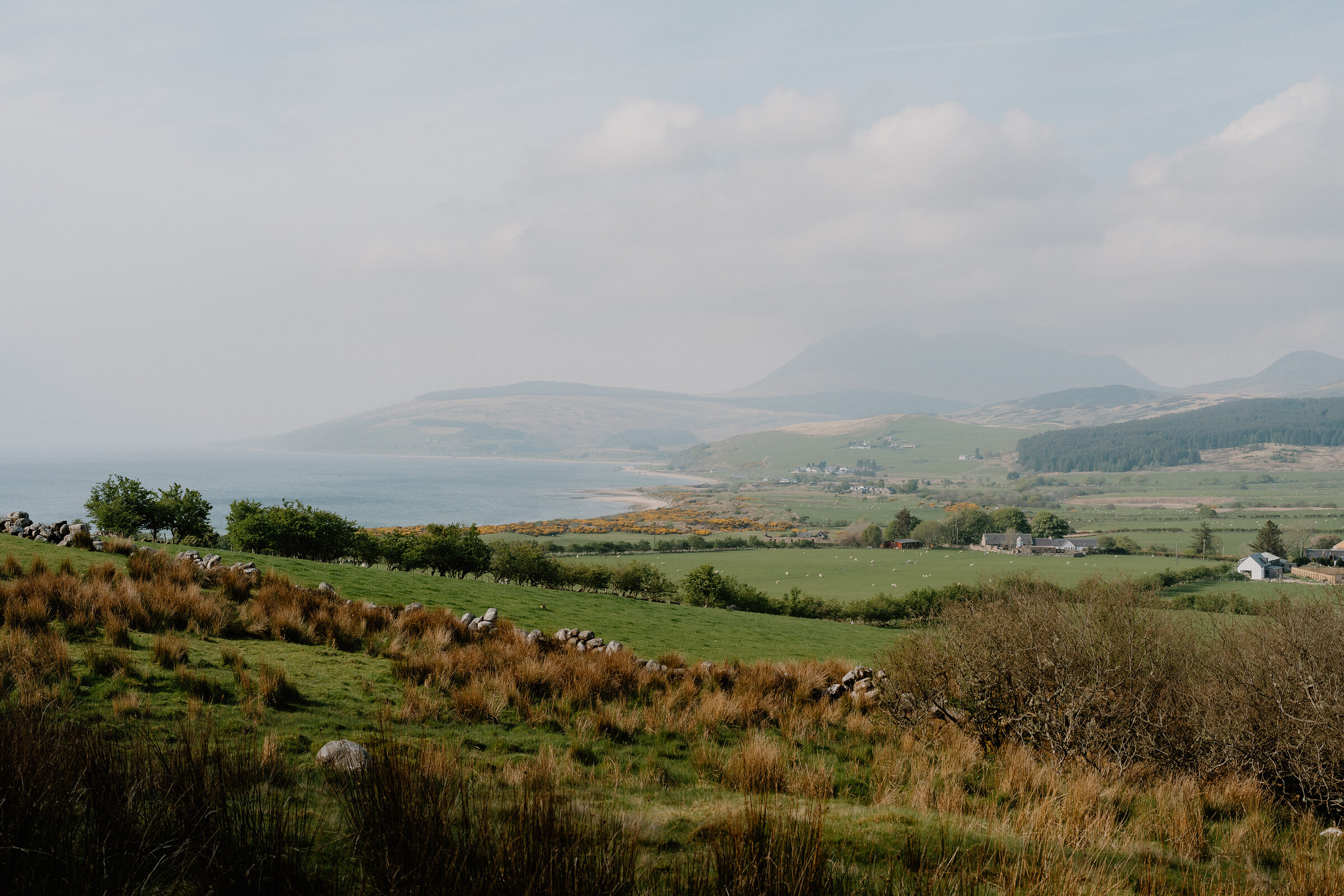 A patchwork of green fields and dry stone walls slopes down to a curved bay, hazy mountains rising across the water.
