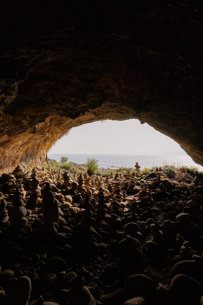 Hundreds of small stacked stone cairns cover the floor of a sea cave, its arched mouth opening onto a view of the sea.
