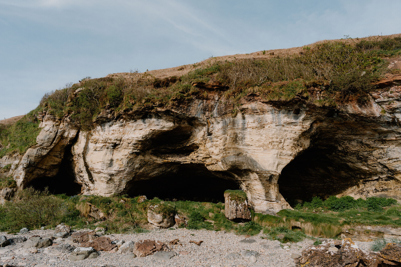 Twin cave mouths open at the base of a scrubby sandstone cliff above a pebble beach.