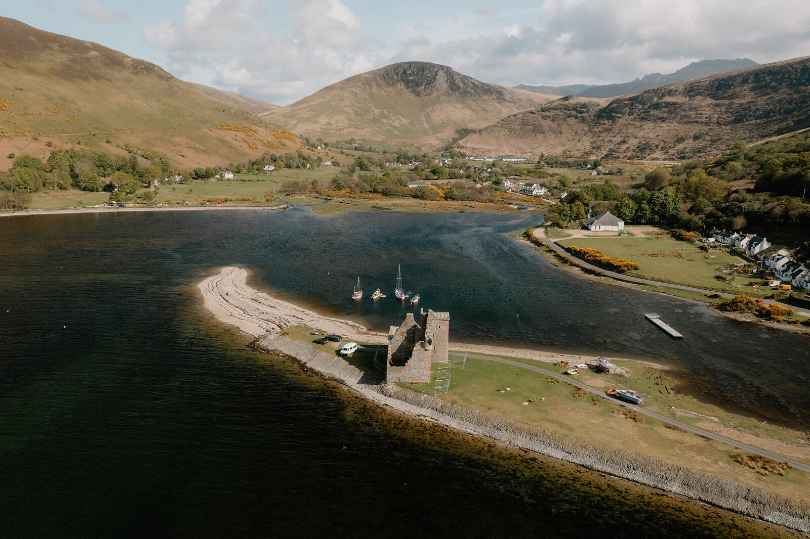 An aerial view shows a ruined castle on a sandy spit reaching into a loch, a village and mountains surrounding the water.