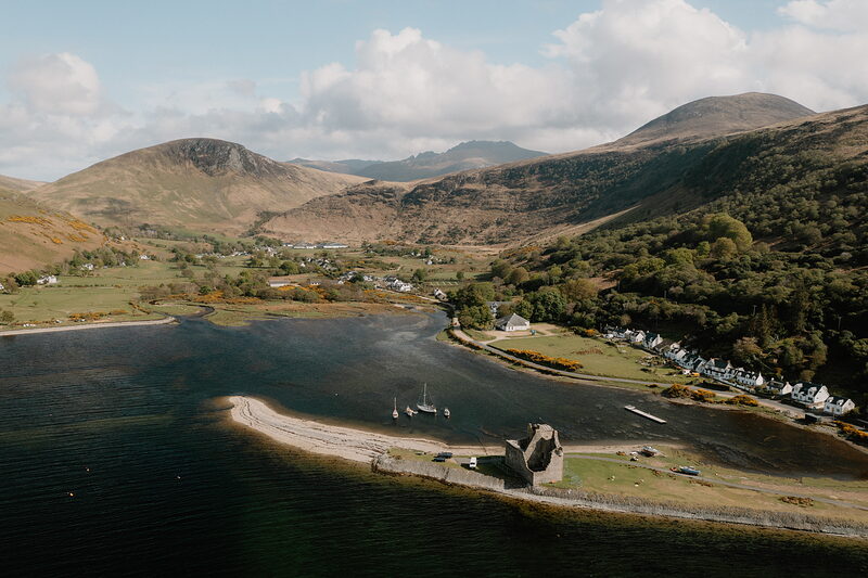 An aerial view shows a ruined castle on a sandy spit reaching into a loch, sailboats moored nearby and a village stretching along the shore.
