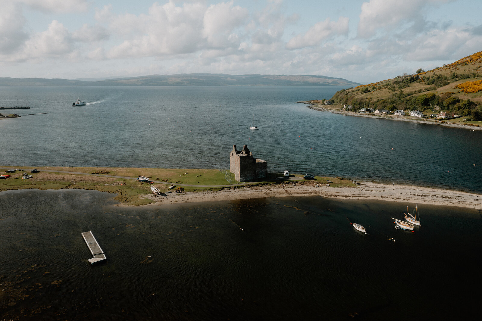 An aerial view shows a ruined castle on a grassy point in a loch, boats moored nearby and hills rising along the shore.
