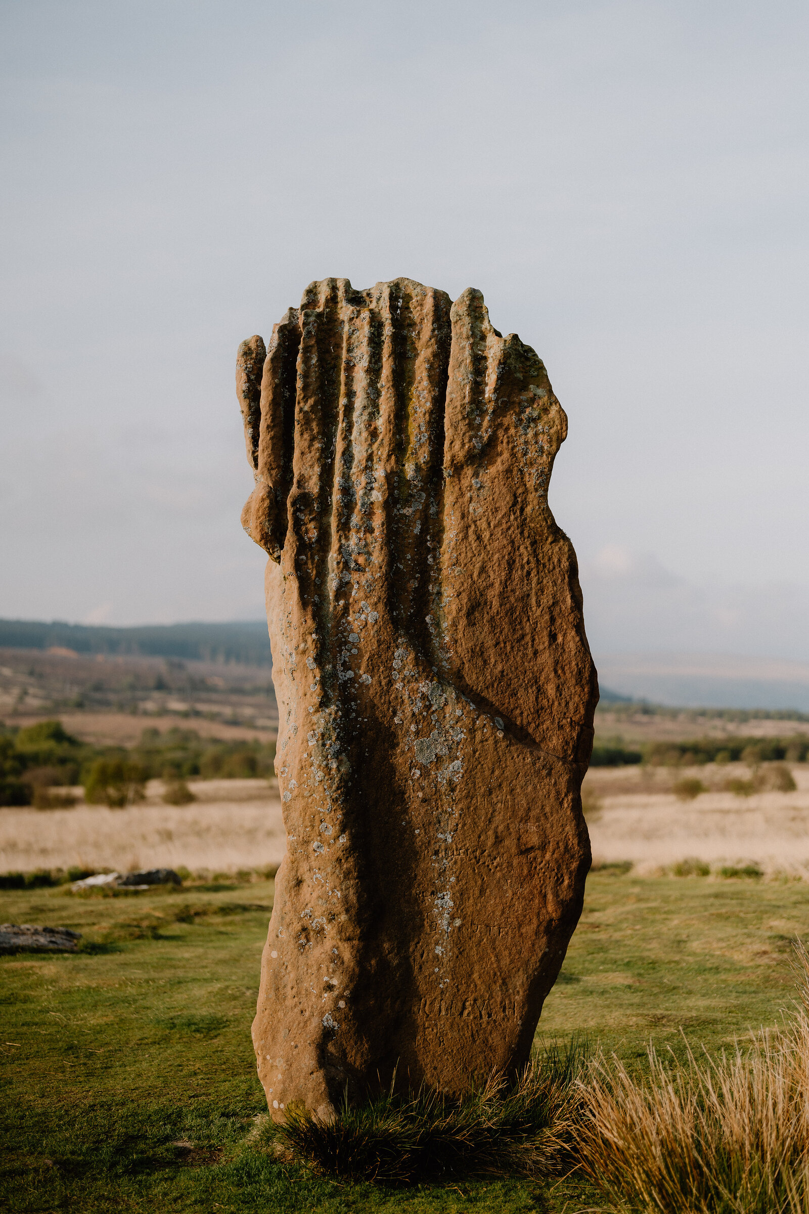 A tall standing stone with deep vertical grooves rises from a grassy field, farmland stretching into the distance.