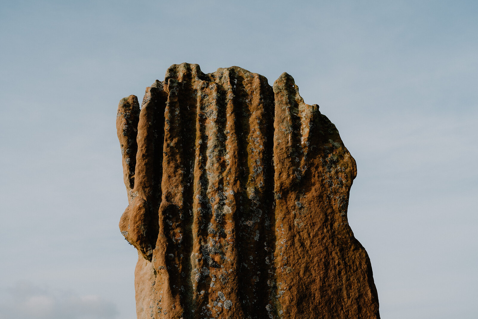 A tall standing stone with deep vertical grooves catches evening light against a pale sky.
