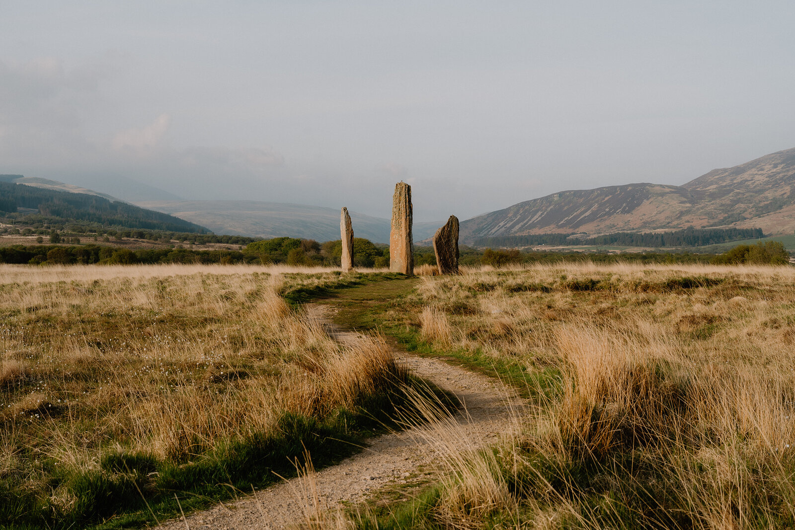 A gravel path curves between three tall standing stones in a grassy field, distant hills fading into haze.