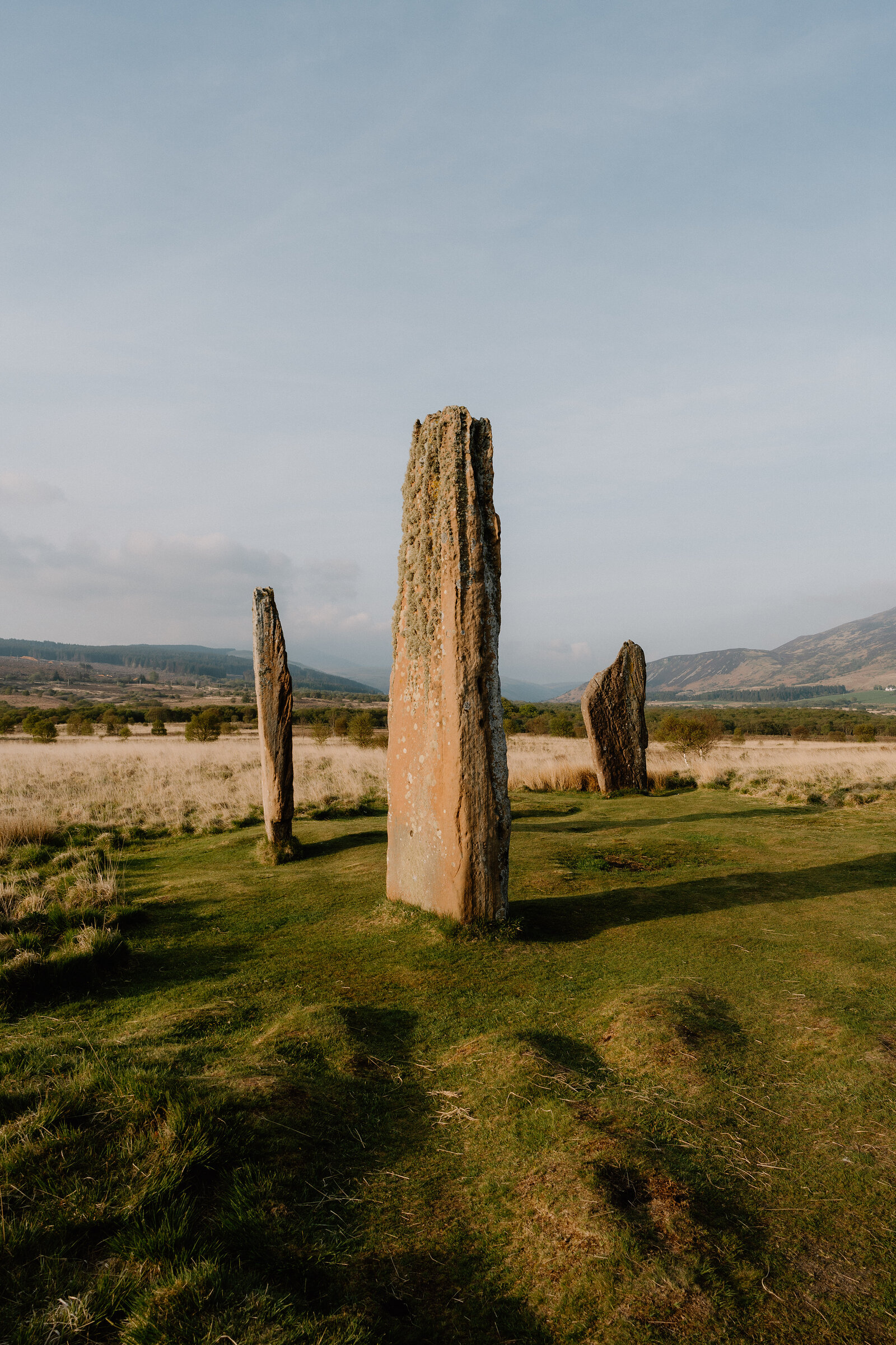 Three tall standing stones rise from a grassy field, hills visible on both sides beneath a clear sky.
