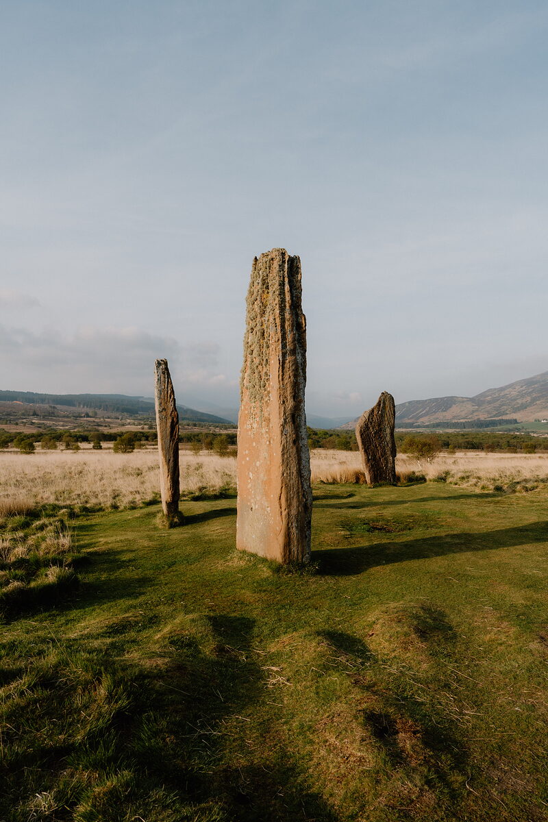 Three tall standing stones rise from a grassy field, hills visible on both sides beneath a clear sky.