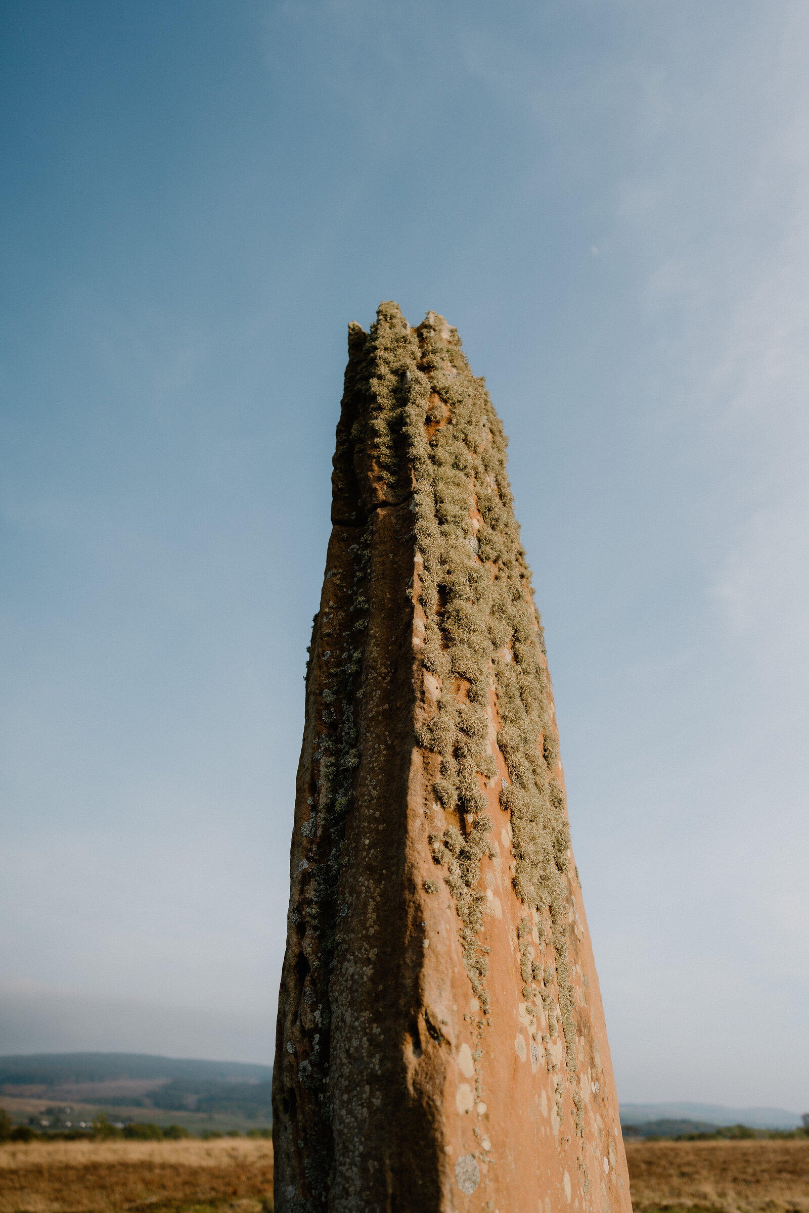 A weathered standing stone streaked with lichen rises against a clear blue sky, distant hills visible at its base.