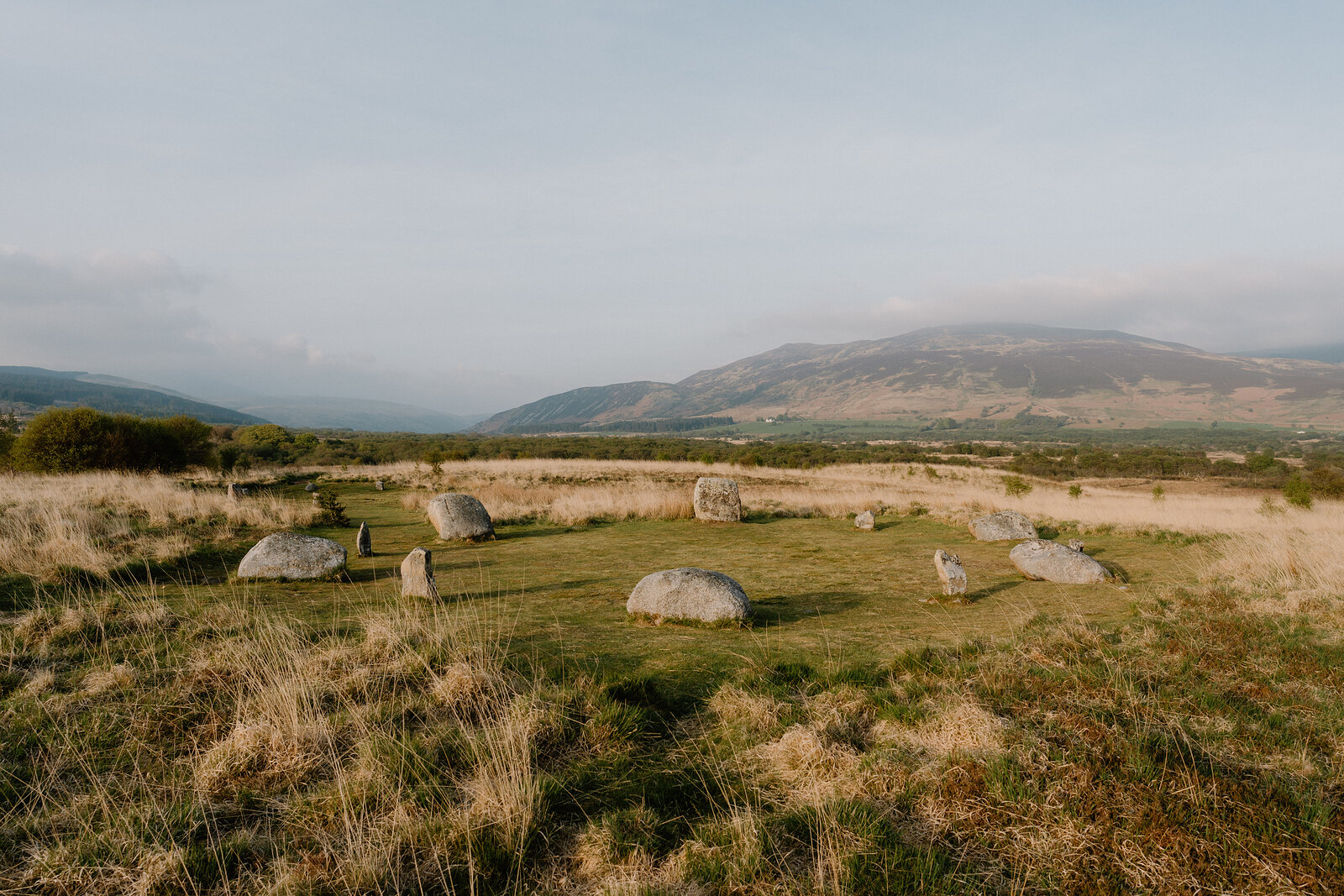Weathered stone hut circles dot a grassy field, wooded hills fading into haze behind.