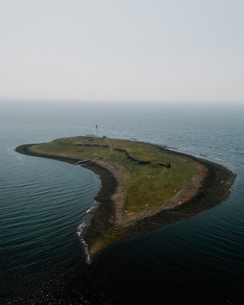 A small tapering rocky island with a lighthouse near its tip lies in open sea, seen from above with hazy sky beyond.