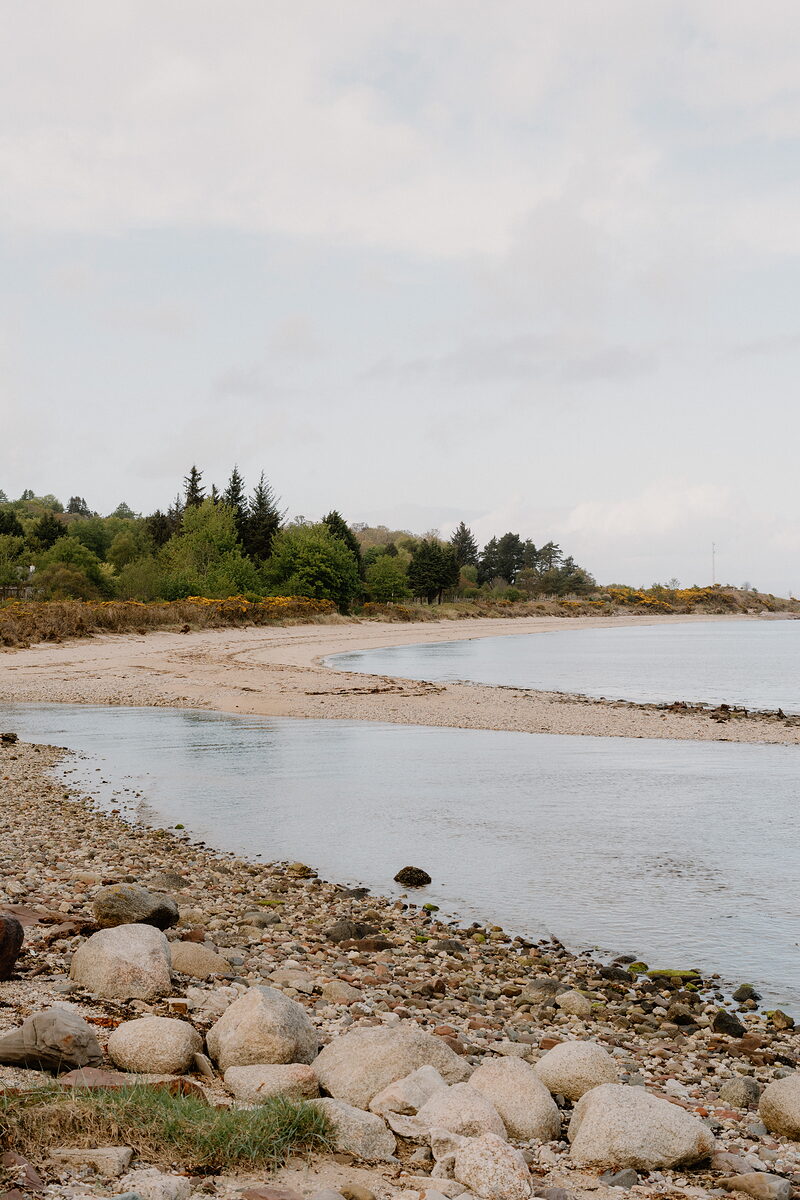A pebble and sand beach curves along a shoreline backed by pine and gorse, with calm sea and a low headland in the distance.