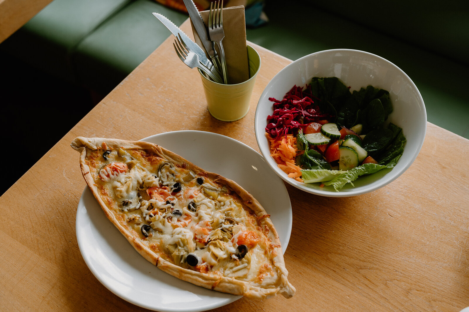A folded savoury pastry topped with melted cheese and olives sits on a plate beside a bowl of mixed salad, a cutlery pot standing on the wooden table.