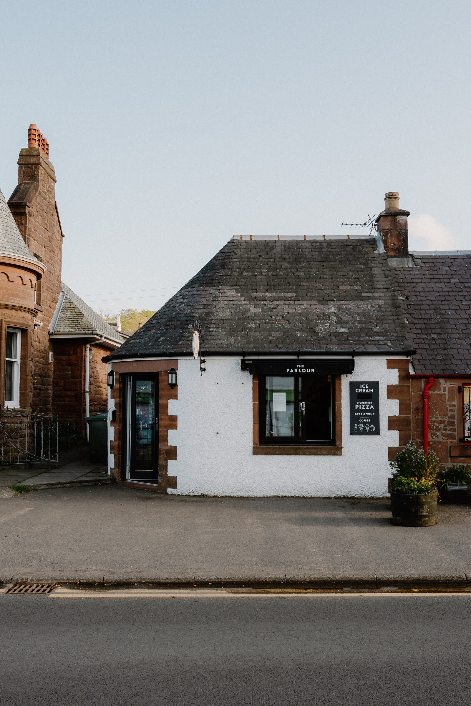 A whitewashed cottage lettered "The Parlour, ice cream, pizza" stands on a street corner beside a sandstone tenement building, its slate roof and chimney visible above.