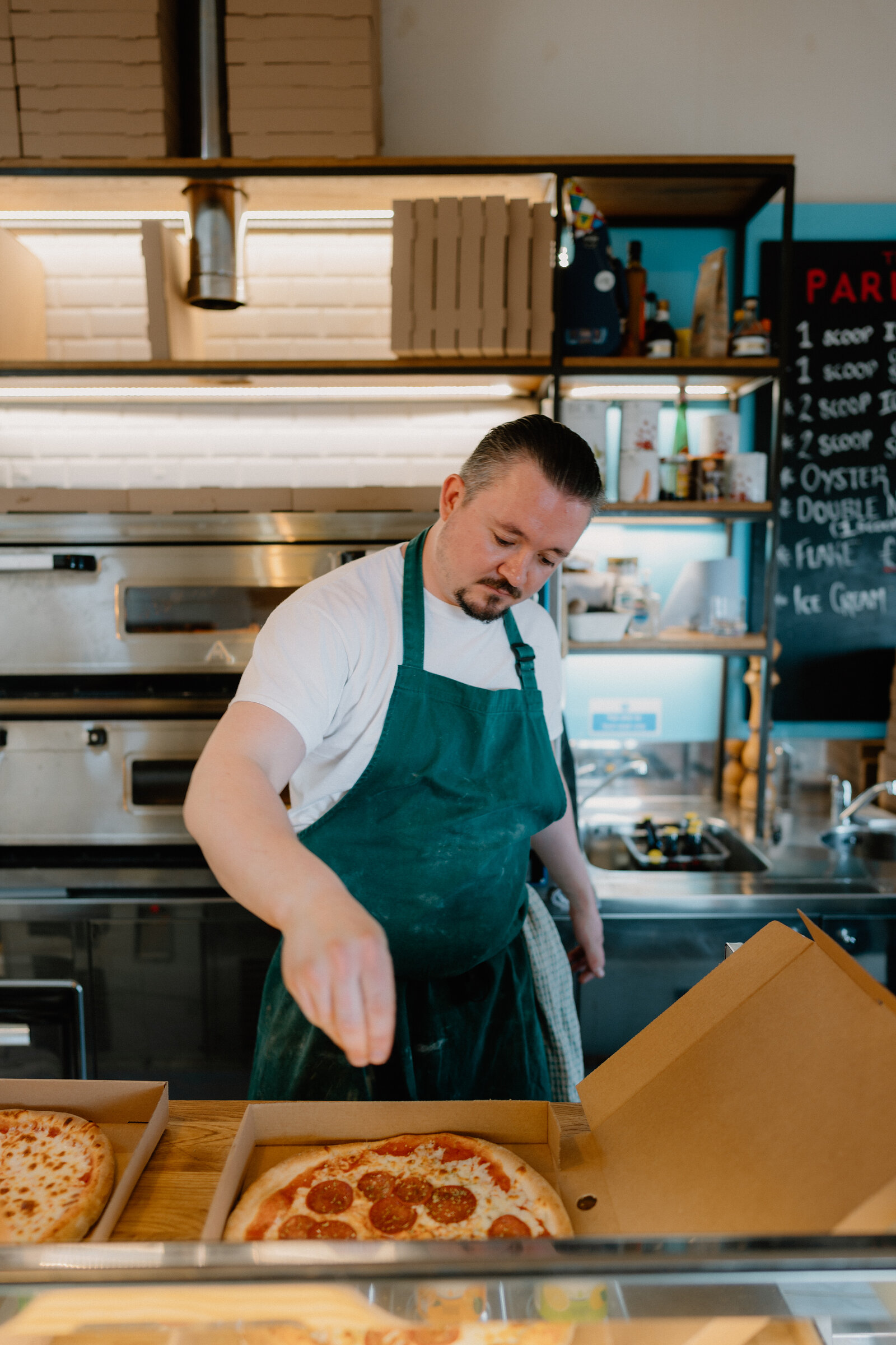 A chef in a green apron places a pepperoni pizza into a takeaway box beside another pizza, shelving of ingredients and a chalkboard menu behind him.