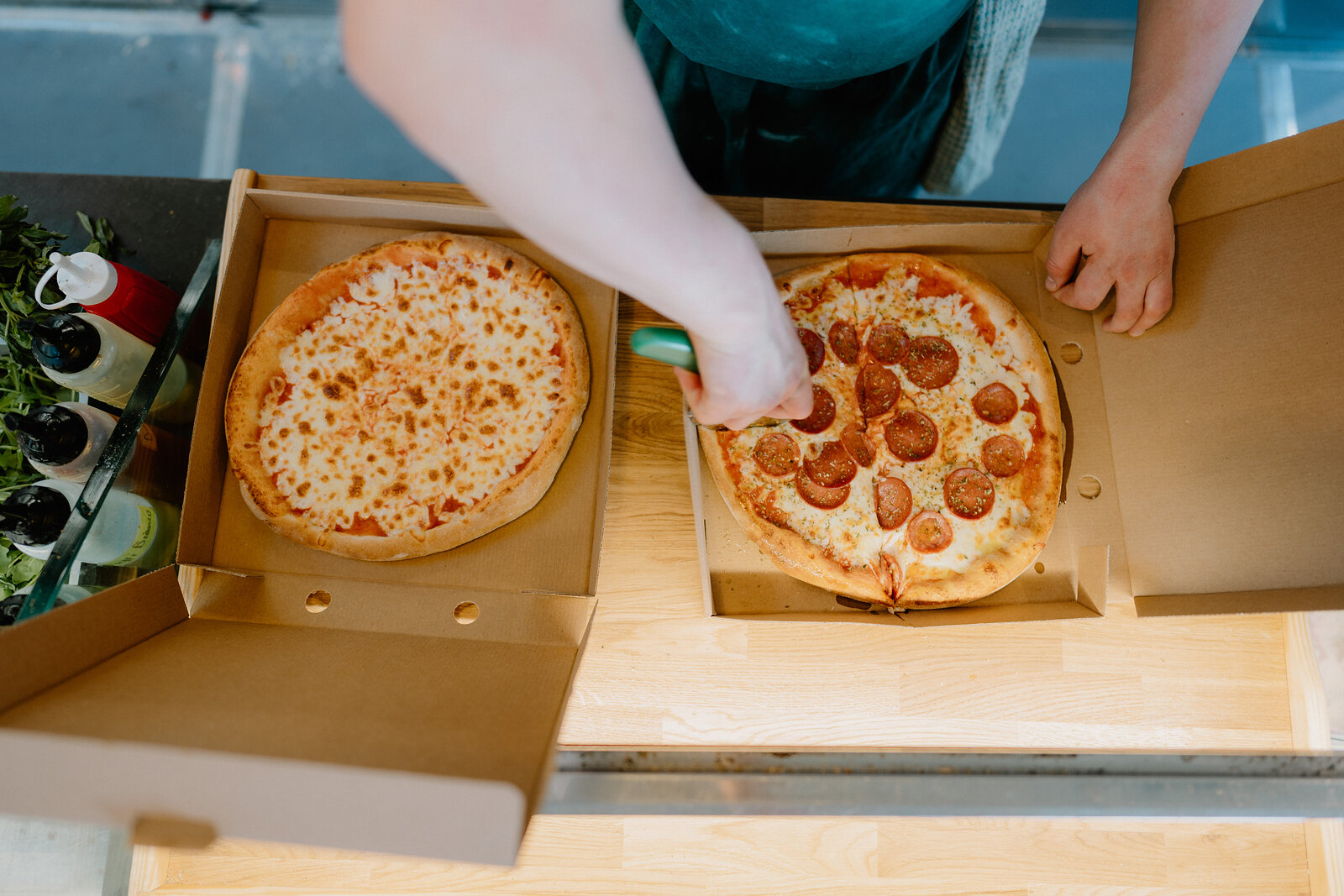 A hand places pepperoni slices onto a cheese pizza beside another topped pizza in open takeaway boxes on a counter.