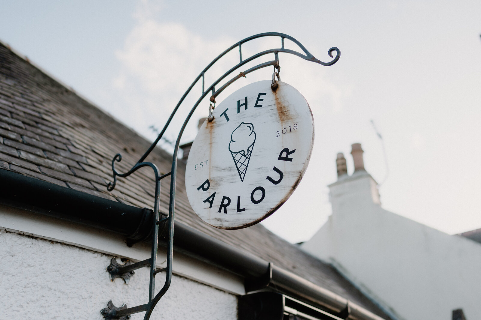 A hanging sign shaped like a wheel reads "The Parlour, est 2018" with an ice cream cone illustration, mounted on an iron bracket above a slate roof.
