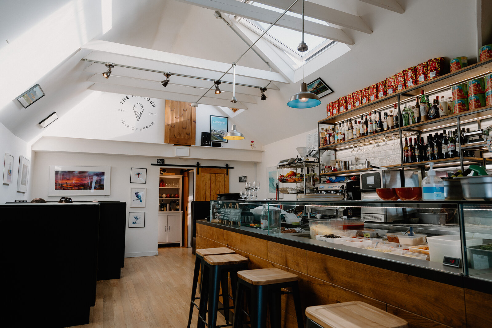 A café with a vaulted ceiling and exposed beams has a wooden counter stocked with tinned tomatoes and drinks, framed prints on the wall and stools lined up in front.