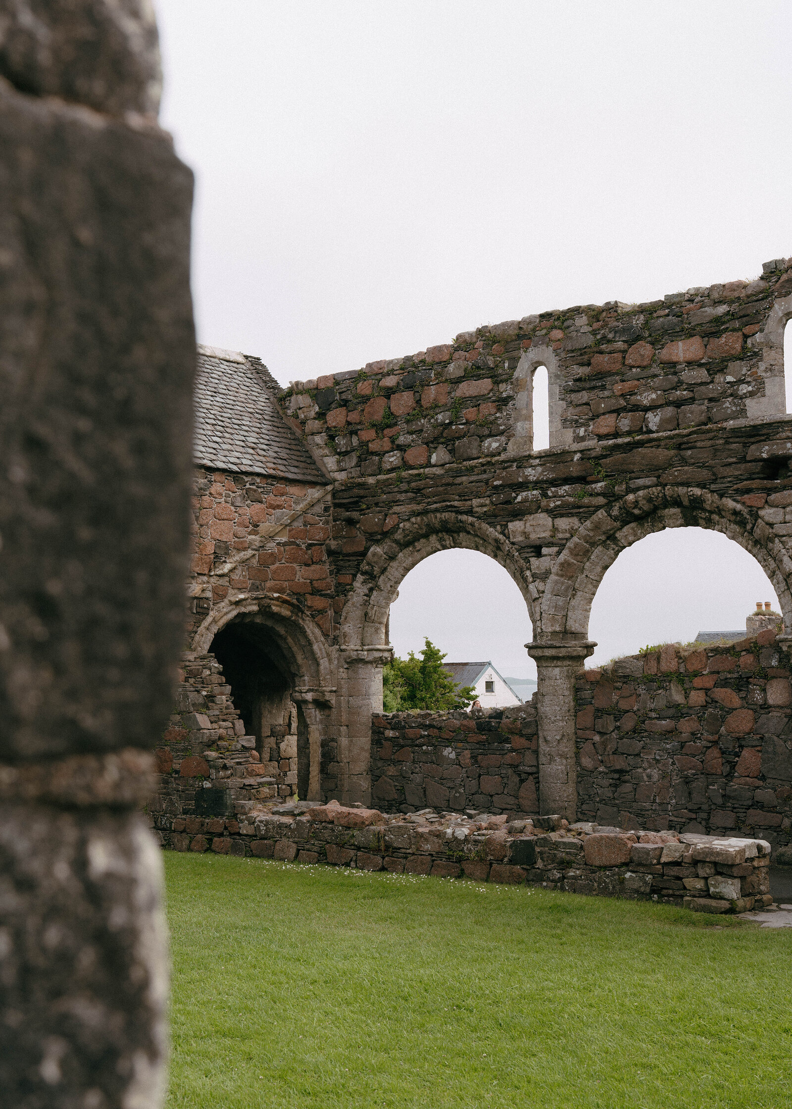 Stone archways of a ruined abbey cloister frame a view of cottage rooftops beyond the grass lawn.
