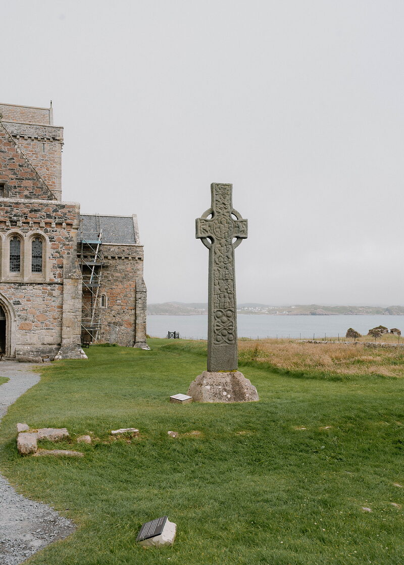 A tall Celtic cross carved with interlace patterns stands on a stone base beside a sandstone abbey, sea and hills visible behind.