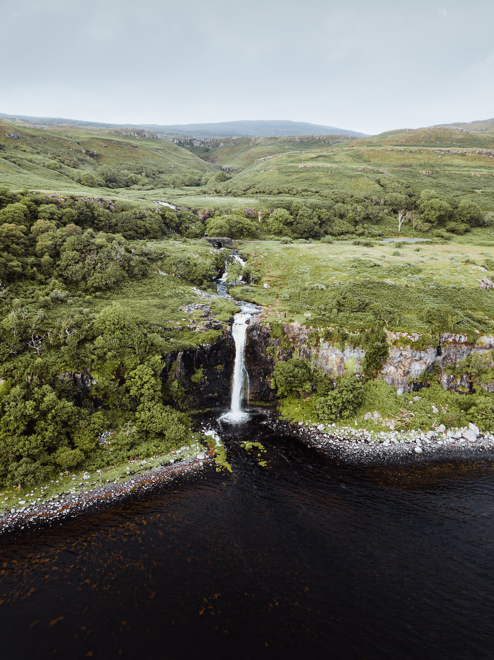 A waterfall tumbles down a rocky cliff into a dark loch, green moorland and woodland spreading out behind.