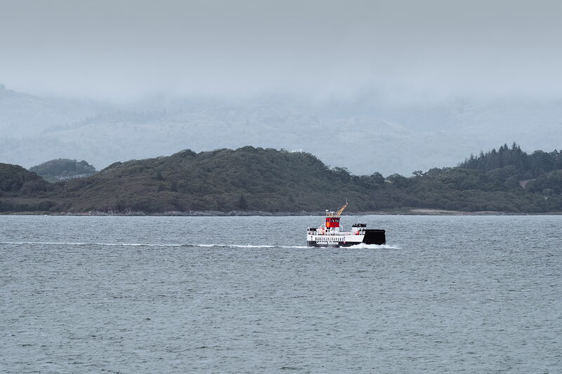A small car ferry crosses calm water, a wooded coastline rising behind it under low cloud.
