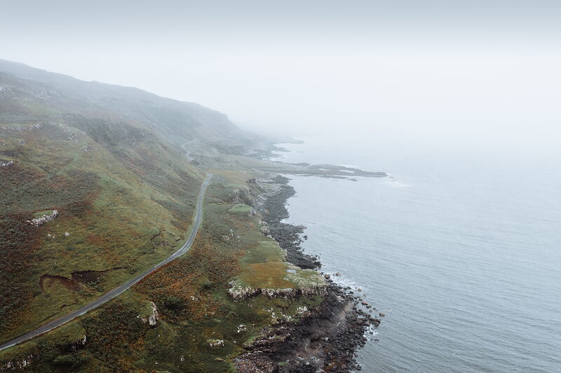 An aerial view follows a coastal road winding along a misty cliff edge, a rocky shoreline stretching below.