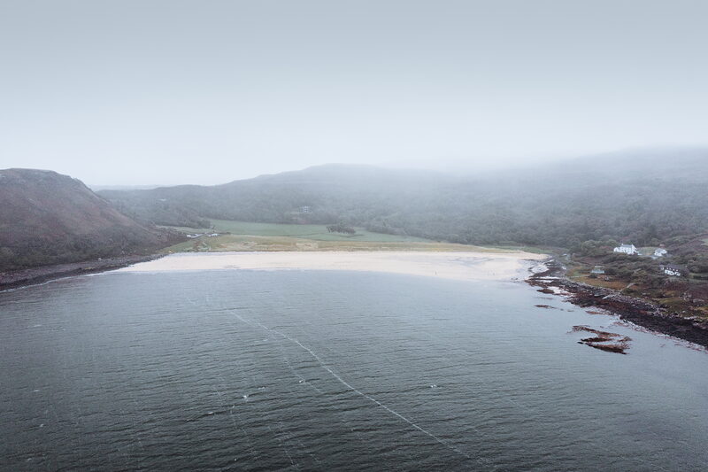 An aerial view shows a misty bay with a sandy beach and green machair, small cottages visible on the far shore.