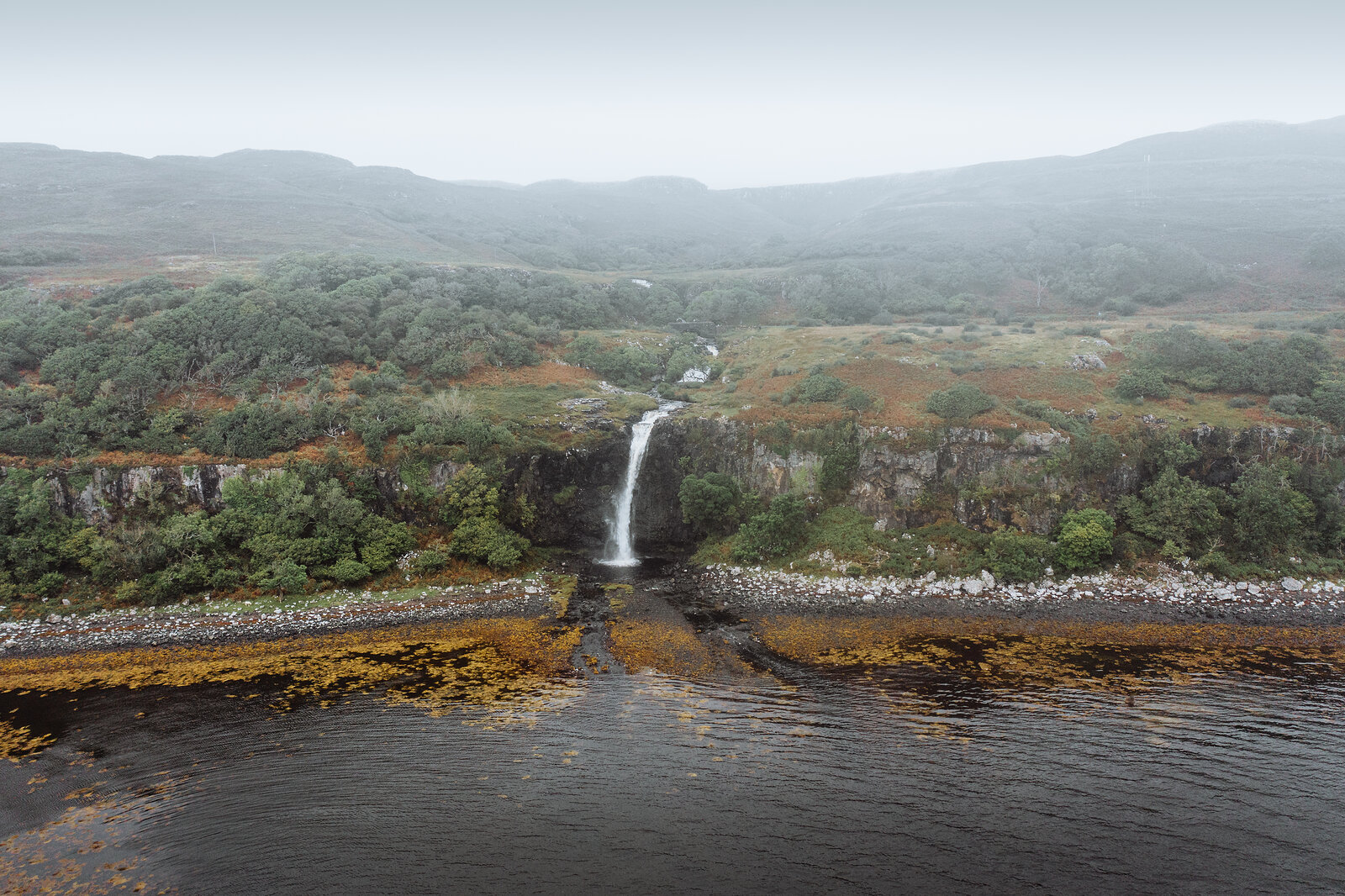An aerial view shows a waterfall dropping into a tidal channel between mossy cliffs, mist softening the hills behind.