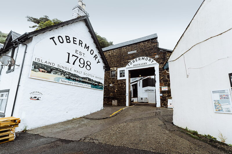 A whitewashed building carries a mural reading "TOBERMORY EST. 1798 ISLAND SINGLE MALT SCOTCH WHISKY", "TOBERMORY DISTILLERY" lettering above a stone archway leading into the yard.
