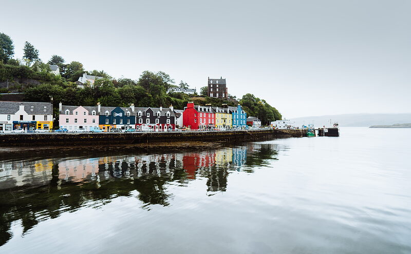 A curved terrace of brightly painted houses lines a harbour, their reflections mirrored in the still water below.