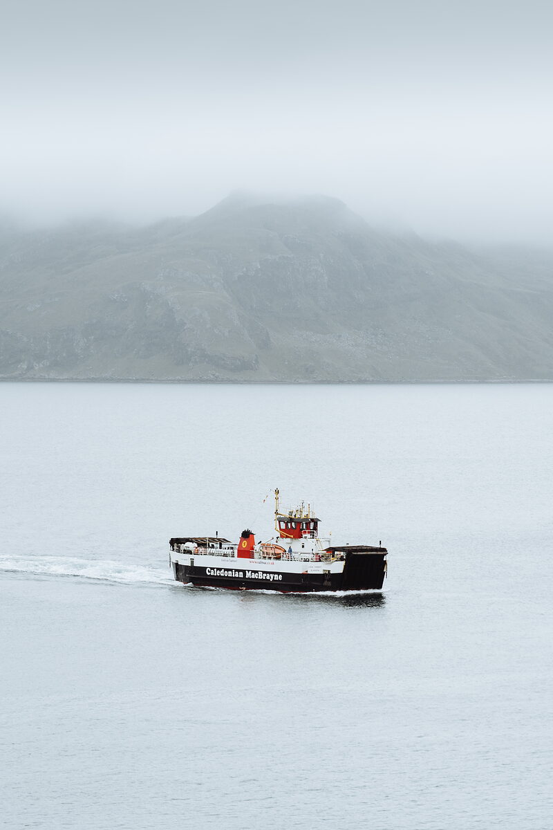 A Caledonian MacBrayne ferry crosses still water beneath a mist-covered mountain.