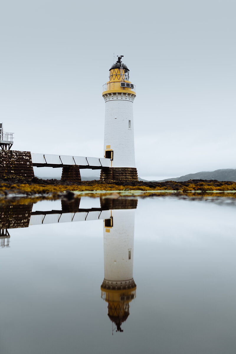 A white lighthouse with a yellow lantern room stands reflected in a still pool, a footbridge and seaweed-covered rocks visible at its base.