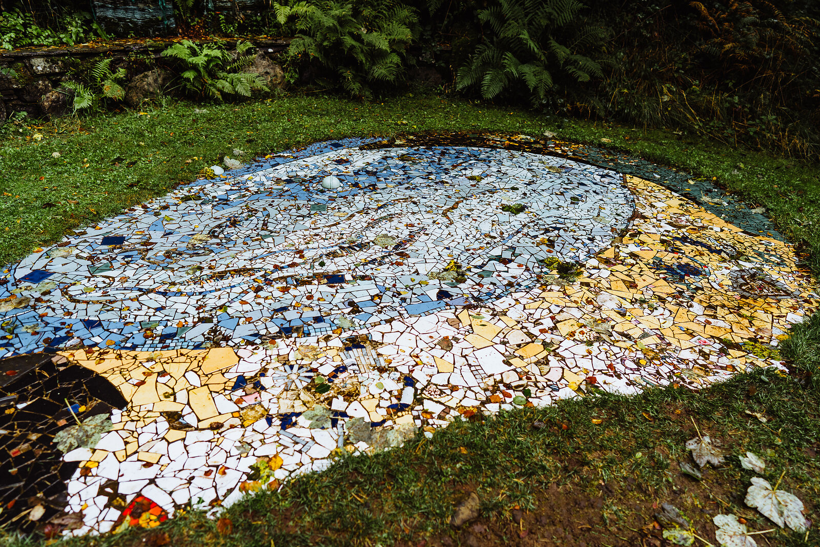 A mosaic made from broken ceramic tiles in blue, white and gold lies set into the grass, part of an outdoor sculpture trail.