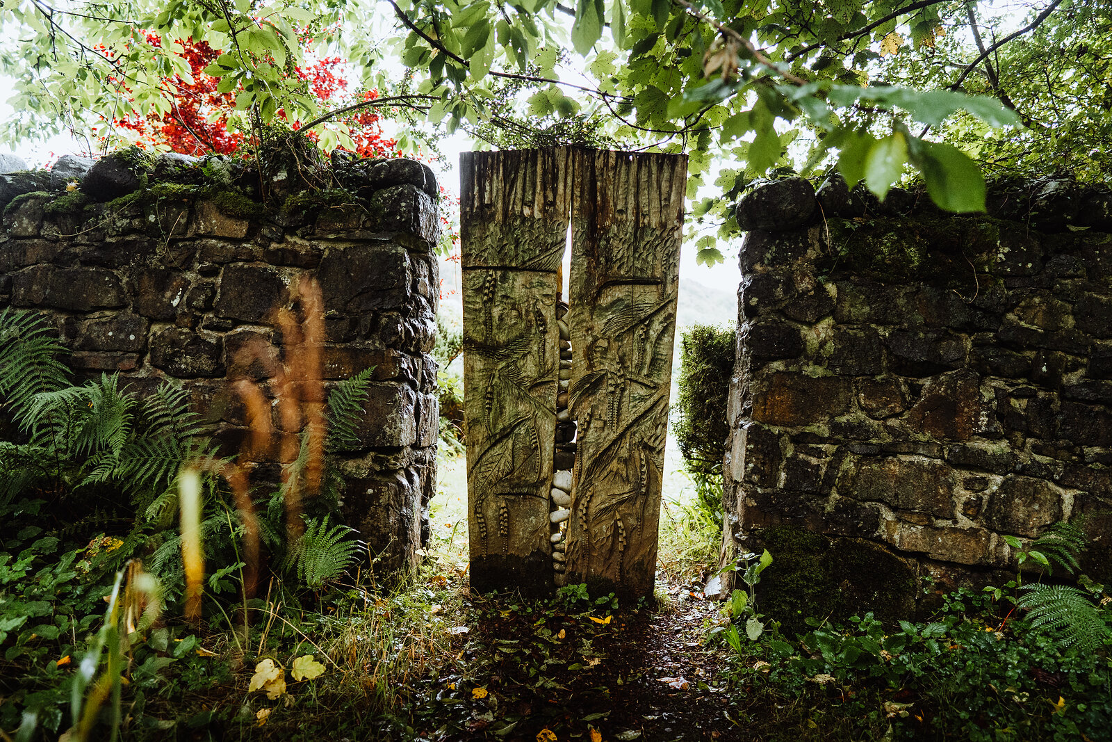 A carved wooden panel depicting a tree stands upright between two mossy stone gateposts, ferns and red flowers growing either side.