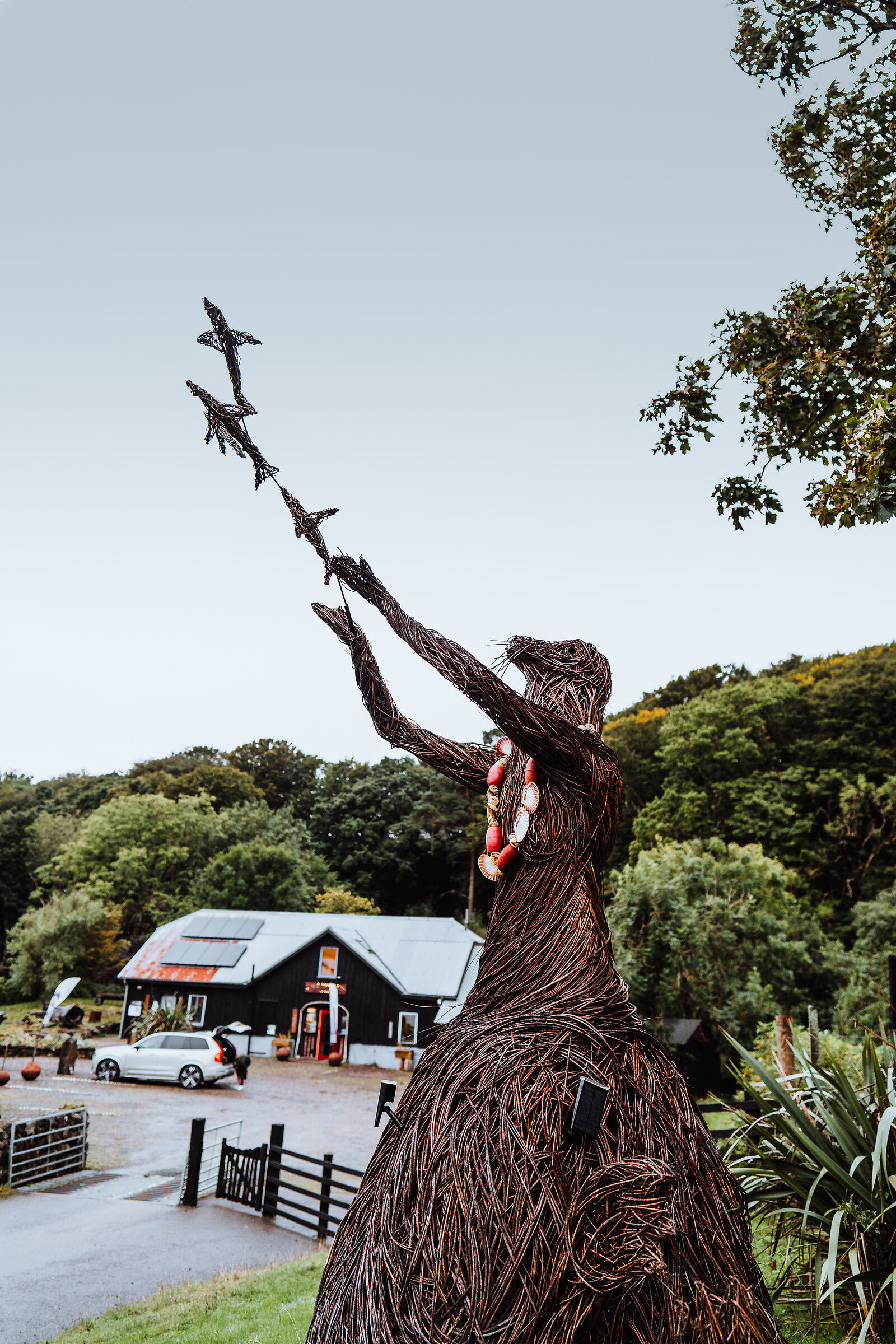 A woven willow sculpture of a robed figure raises two birds skyward, a farm shop and parked car visible in the field behind.