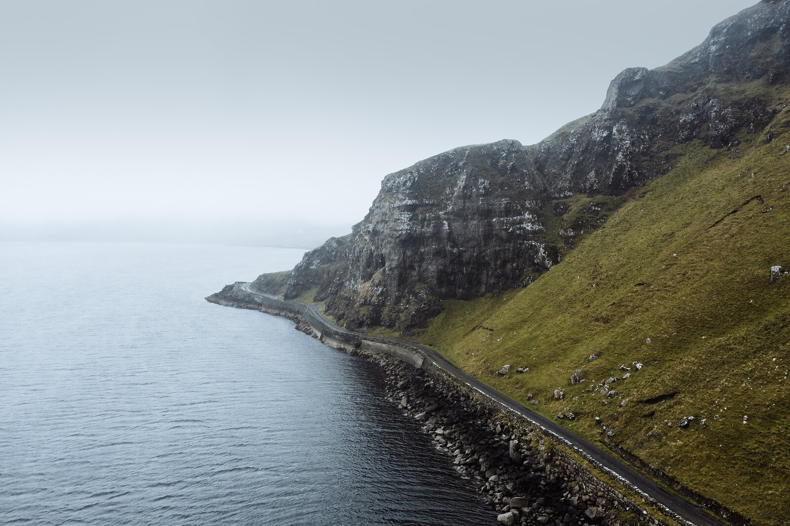 A single-track road hugs the base of steep, grassy cliffs above a calm sea shrouded in mist.