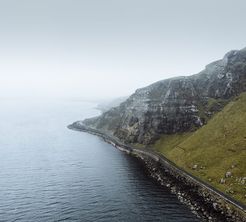 A single-track road hugs the base of steep, layered cliffs above a misty sea.