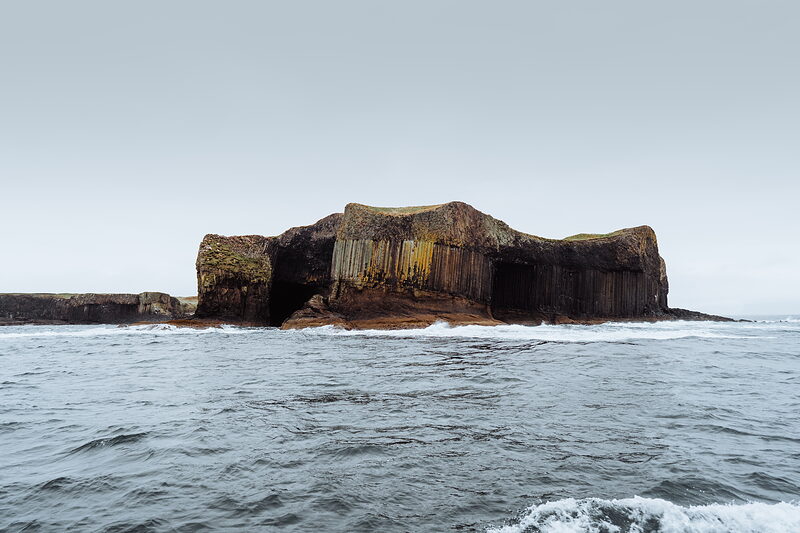 A rock islet riddled with columned basalt caves and pillars rises from a calm sea.