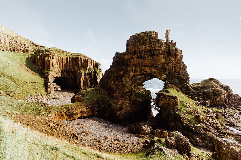 A tall rock arch rises from a rugged coastal outcrop with layered basalt cliffs on one side, the sea visible through the opening.