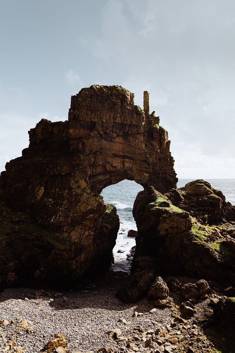 A tall natural rock arch rises from a pebble beach, the sea visible through its opening.