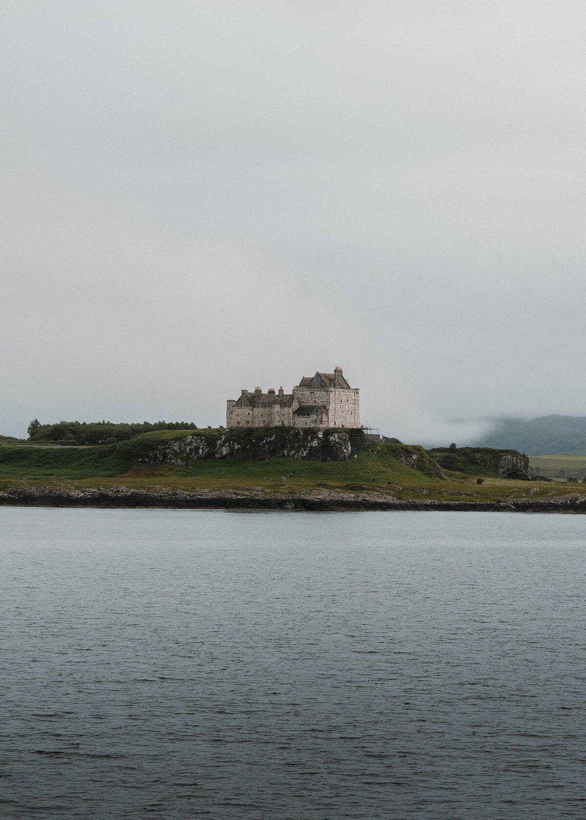 An upright frame of Duart Castle on its headland, a wide band of still grey water beneath and mist behind.