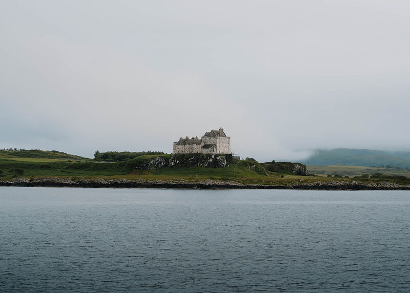 Duart Castle closer, its pale walls and crow-stepped gables above the rocks, green fields and misty hills behind.