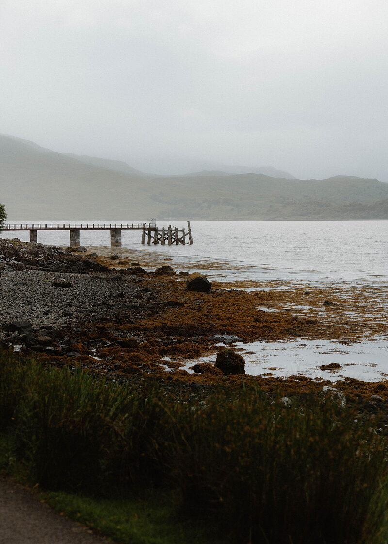 A stony shore of orange seaweed and rocks, an old wooden jetty and misty hills across the loch behind.