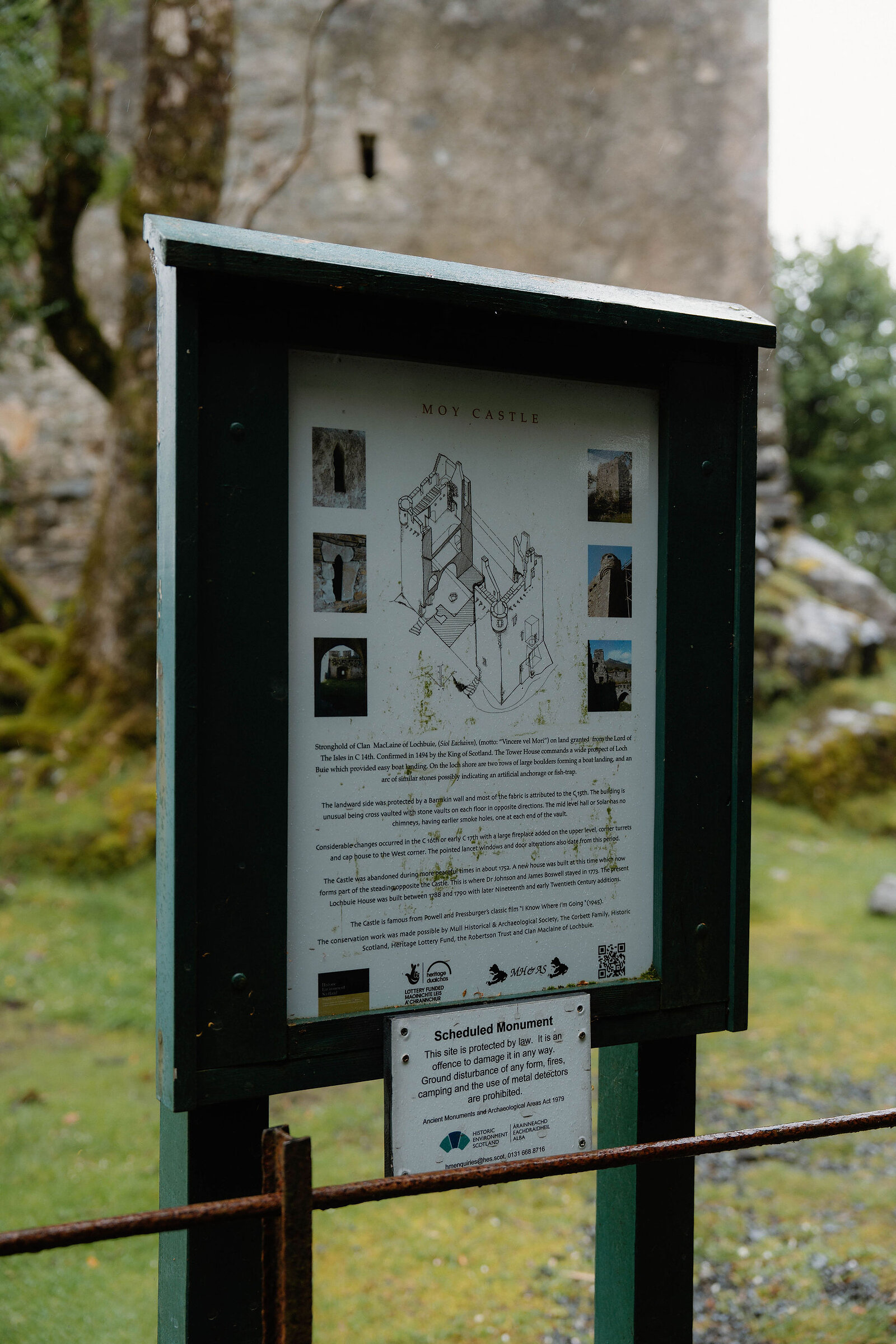 A green-framed information board for Moy Castle on posts by a rail, the tower rising behind.
