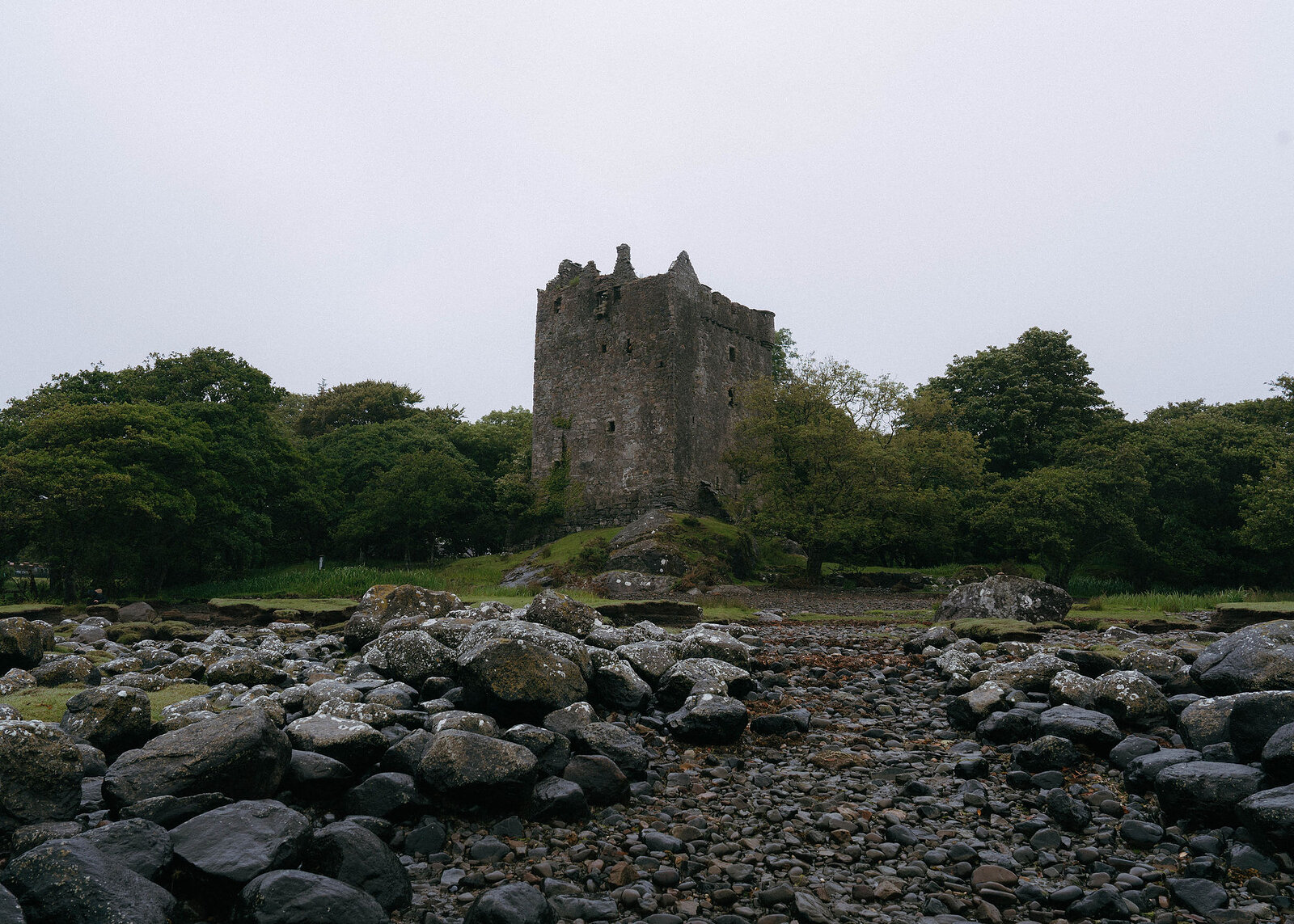 Moy Castle framed by trees, a channel of pebbles running up to it between banks of grey boulders.