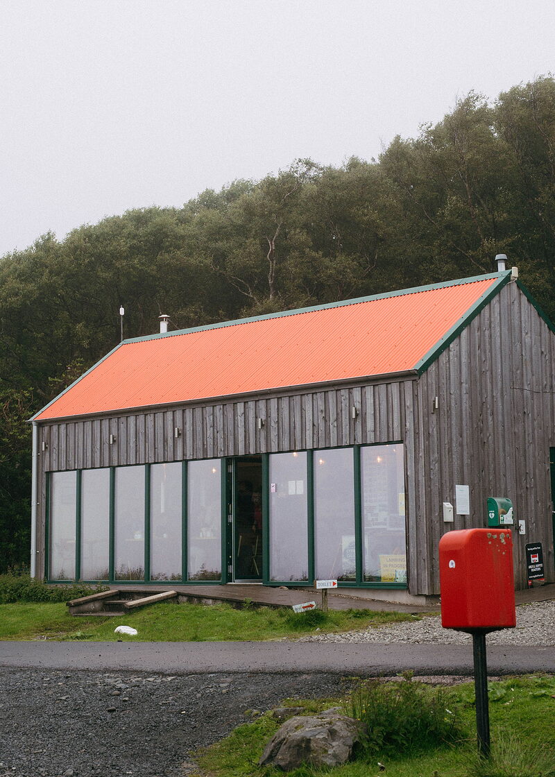 A timber-clad shop with a bright orange roof and tall windows, a red postbox by the road and trees behind.