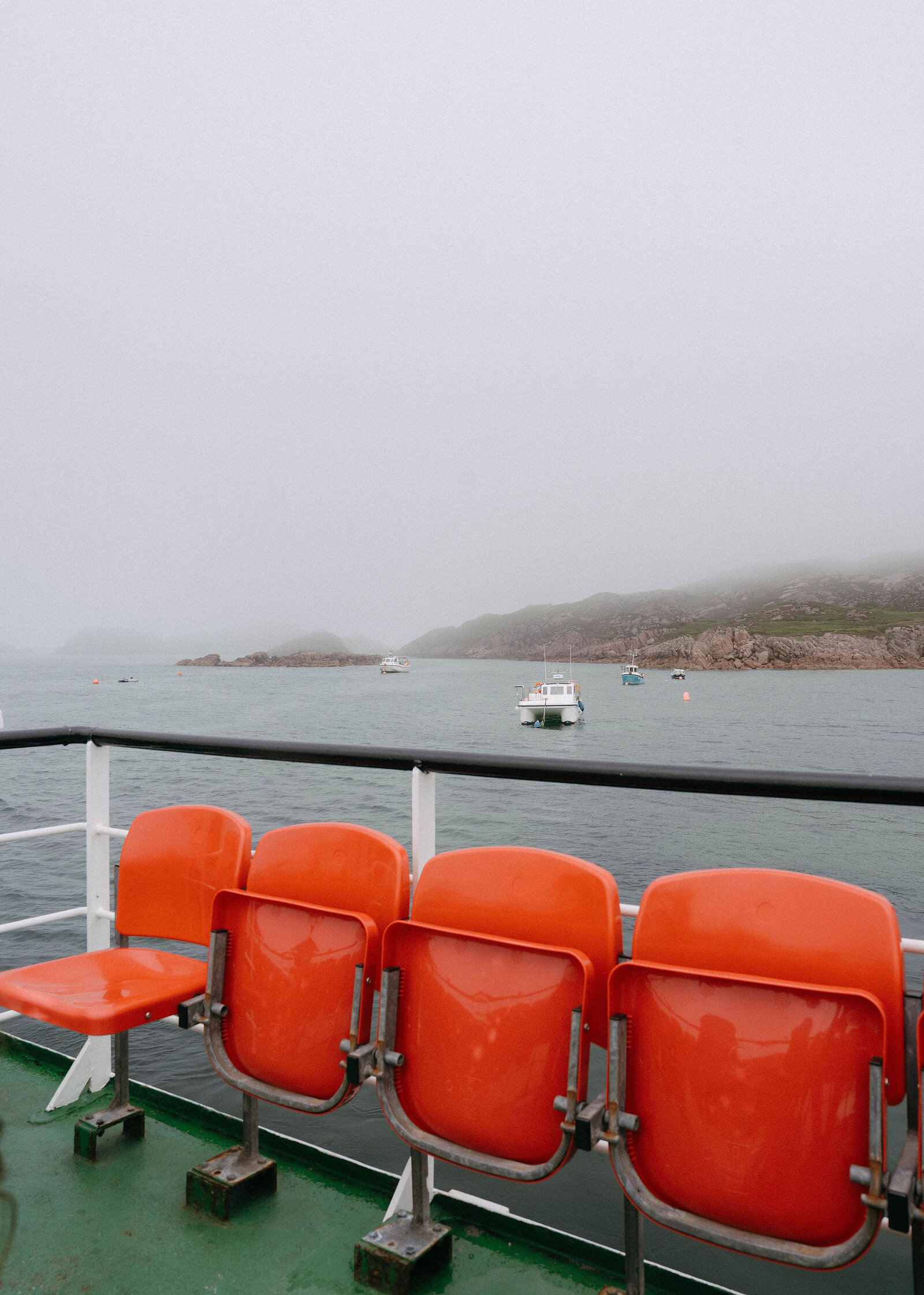 A row of orange seats on the ferry deck, small boats moored off a rocky shore under low mist.
