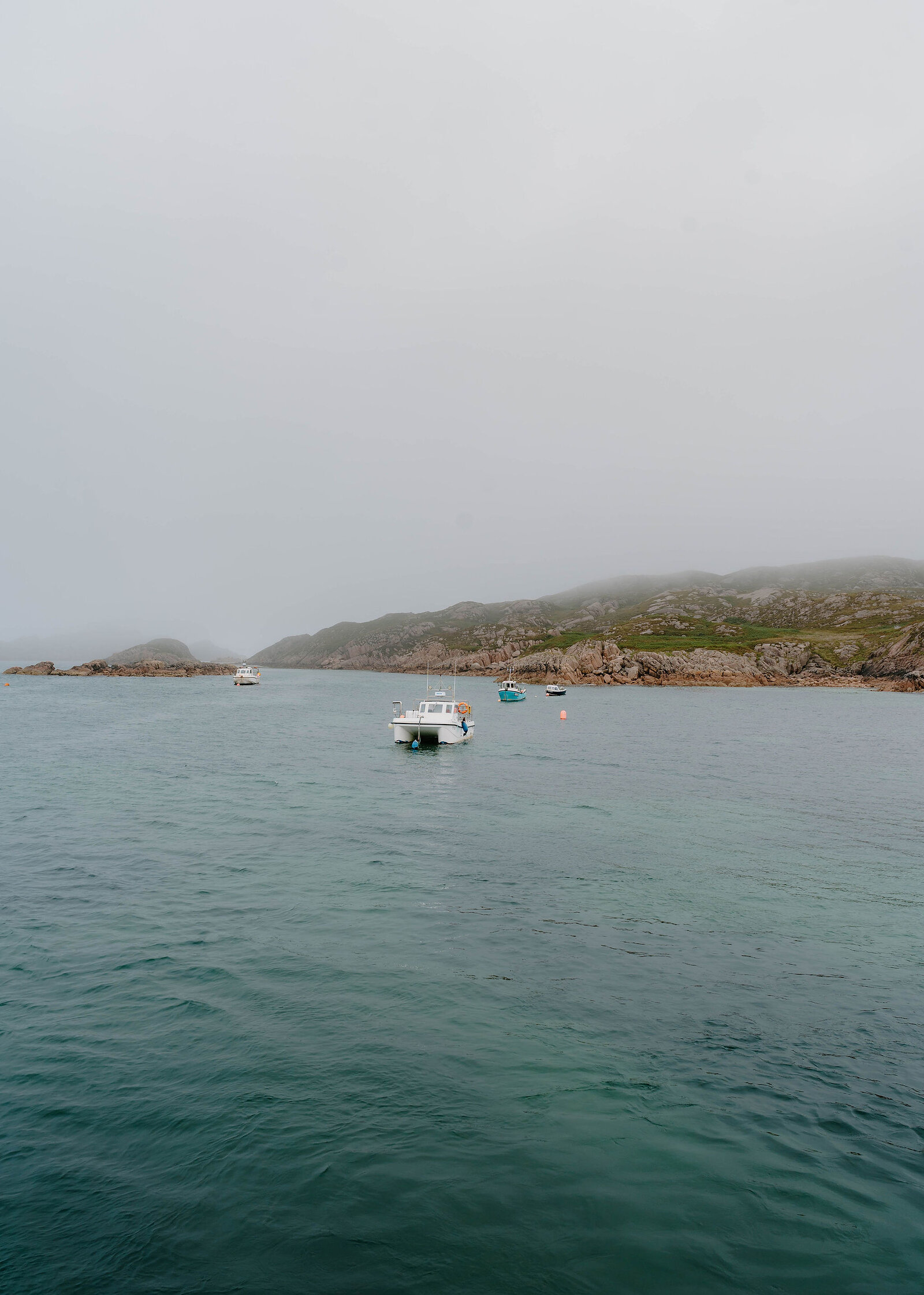 Green water and small boats moored in the sound, pink granite rocks along the misty far shore.