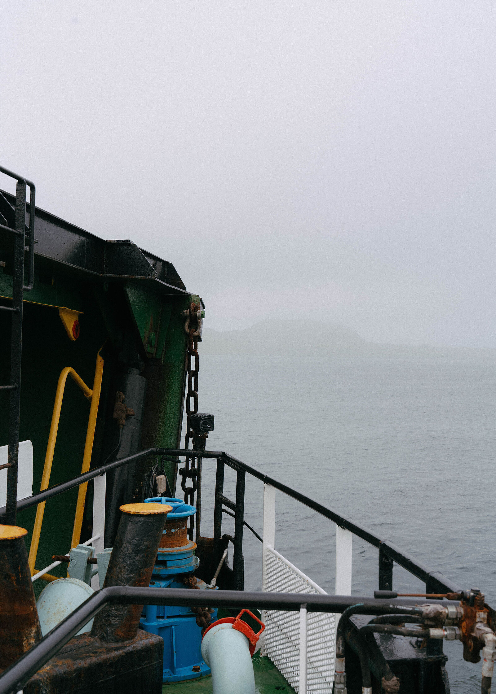 The ferry's green side, ramp chains and blue winch on the right, grey sea and a faint island ahead.
